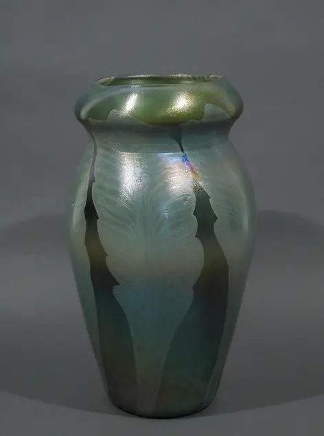 Green vase against a grey background. Two large greenish grey leaves are visible on the vase. The leaves appear lighter than the background. Light reflects off the upper third of the vase.