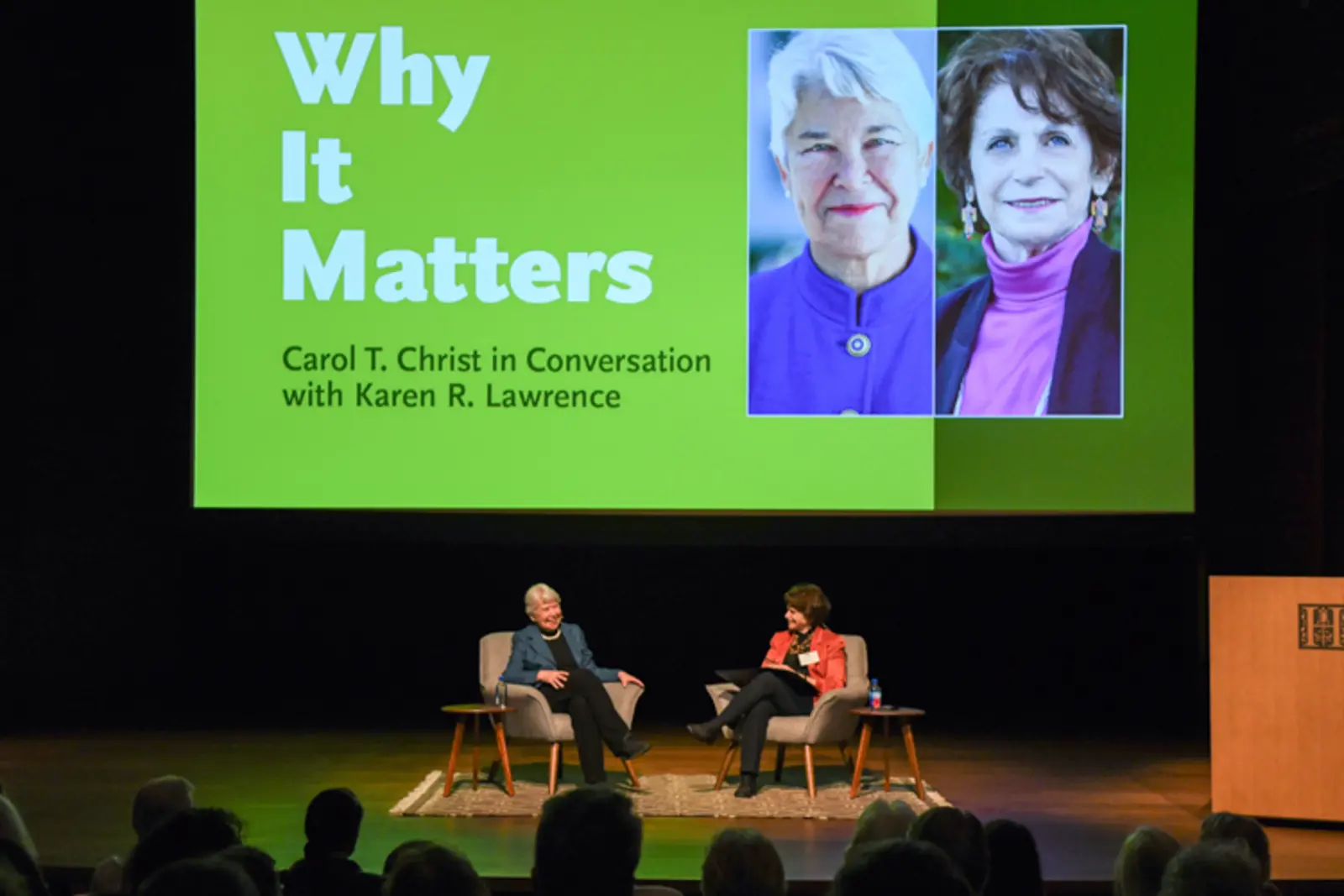 Two people sit in chairs on a stage in front of an audience. A large projection screen has two portraits and reads “Why It Matters: Carol T. Christ in Conversation with Karen R. Lawrence.”